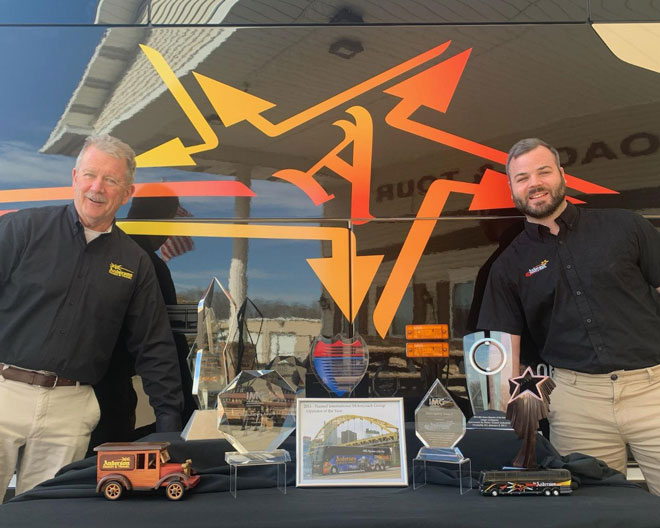 Doug and Evan Anderson with table of awards in front of bus